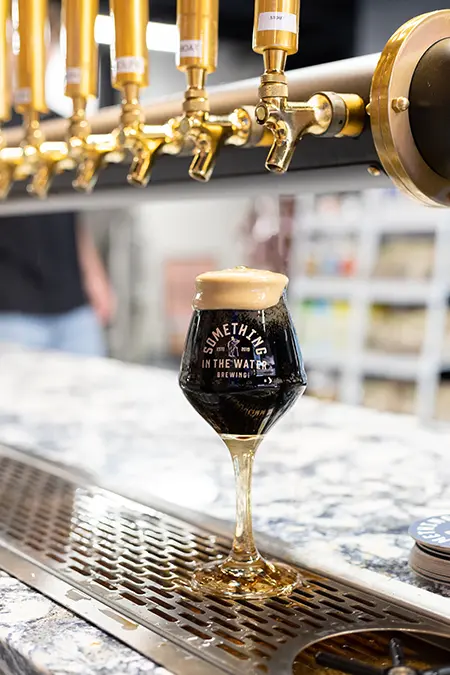 Dark beer with a foam head in a stemmed glass beneath brass taps at the Something in the Water taproom